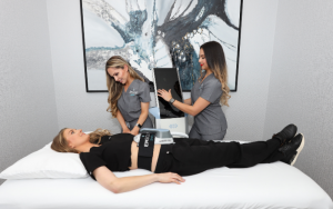 Two women in gray scrubs helping apply emsculpt machine to stomach